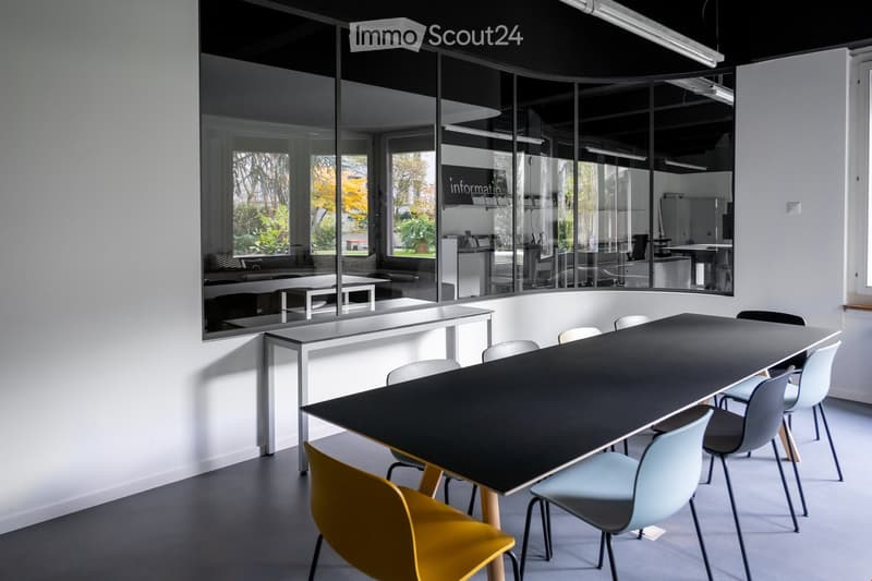 Conference room with white walls, glass walls, gray floor, a large long black table, colorful chairs, and other furniture