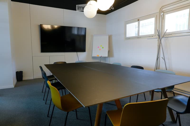 A modern meeting room with a large rectangular table surrounded by colorful chairs, a mounted screen, a whiteboard with sticky notes, and a hanging lamp.