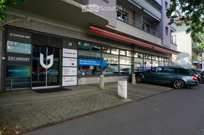 Modern commercial building with glass storefronts, signage for 'Backup One' and 'Dreamhunt Studio', and a parked car in front.