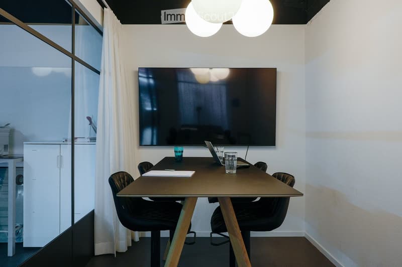 Modern office space with a large screen, a wooden table, chairs, and a laptop. The room has large windows with white curtains and modern lighting.