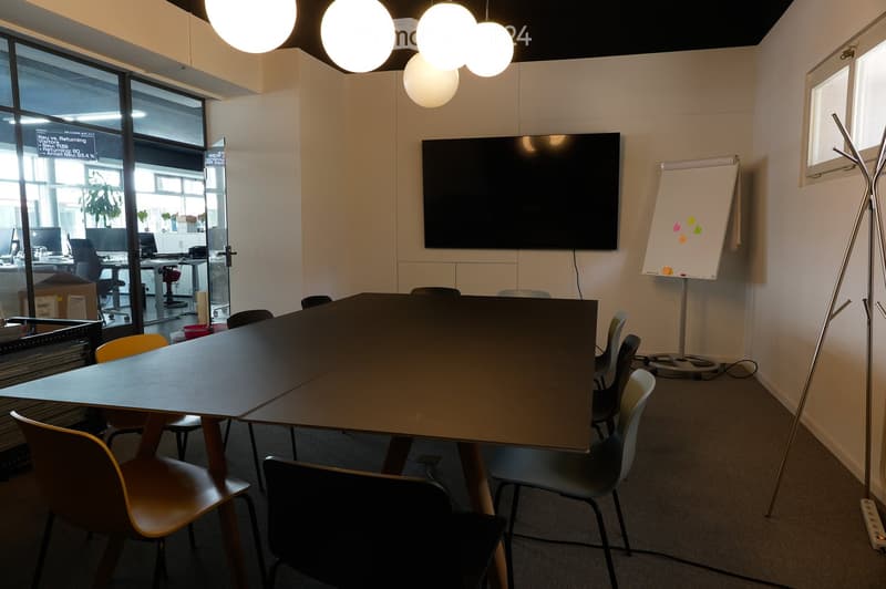 Modern office meeting room with a large table, chairs, a large screen, and a screen stand. The room has large windows and modern lighting.