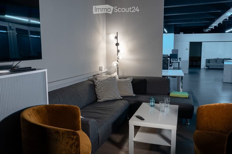 Modern living room with a grey sectional sofa, patterned throw pillows, a white coffee table with glasses and a remote, and a sleek screen mounted on the wall. The room features a warm, inviting ambiance with contemporary decor and additional seating in t