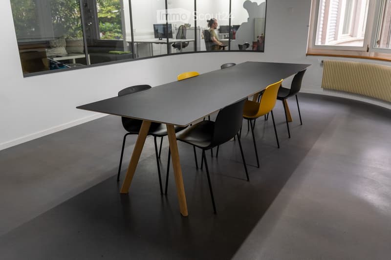 Modern office space with a long black table and several chairs, large windows with a view of greenery, and a radiator on the wall.