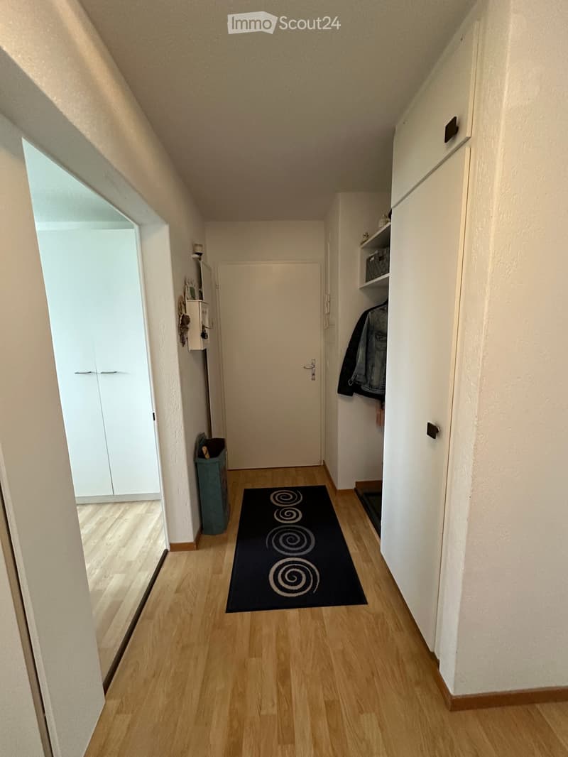 Corridor with wooden floors, two white doors, one open leading to another room, a trash bin, a black patterned rug, and a closet with a basket.