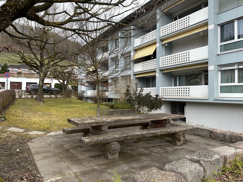 Garden with wooden picnic table and bench. Grassy area in front of an apartment building with balconies.