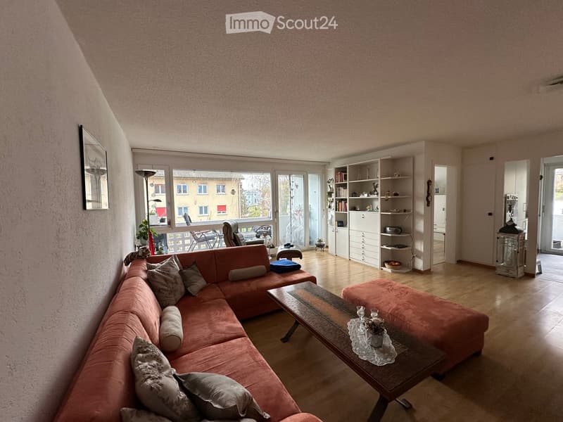 Modern apartment with a spacious living room. Features an L-shaped orange sofa with pillows, a center table, and an armchair. There is a balcony with a view of the street. The room has white walls, wooden floors, and white ceiling. There is a cabinet with