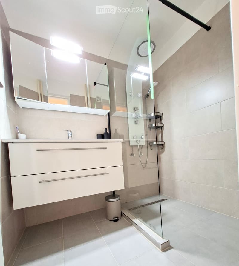 Modern bathroom with gray tiles, glass shower enclosure, white vanity with drawers, mirror with lights, and a silver trash can