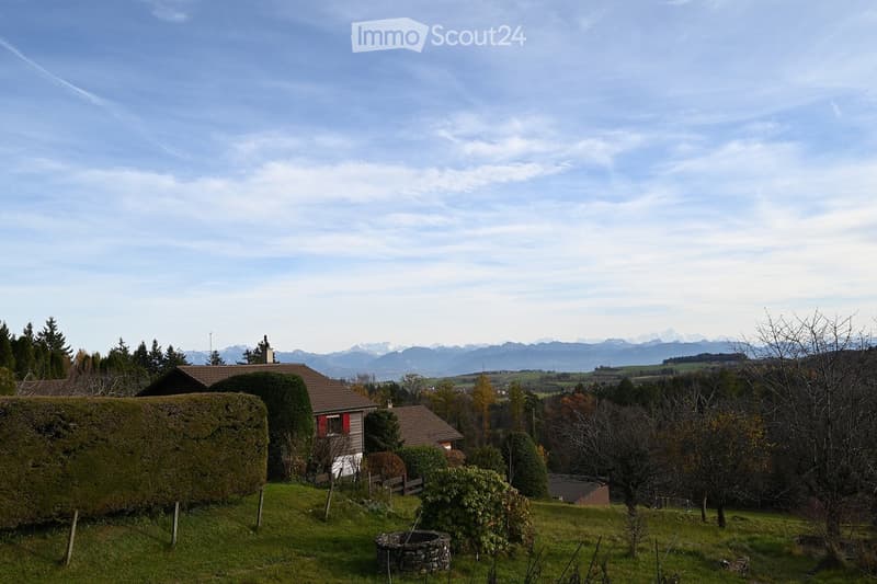 Detached house, two stories, red roof, red shutters, surrounded by trees, open area, grassy garden, trees and bushes, distant mountain range