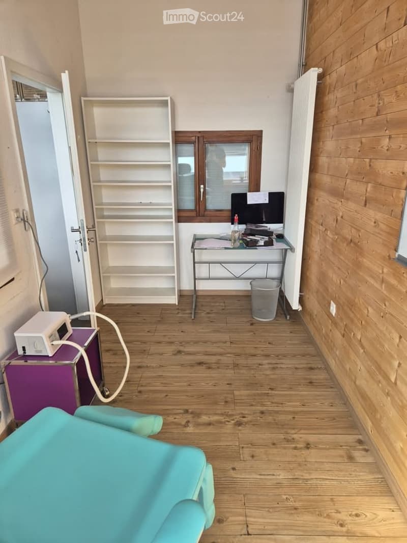 An empty room with wooden floor, white walls and wooden wall, a desk, a chair, a shelf and a trash can, a machine on a table, a massage table.