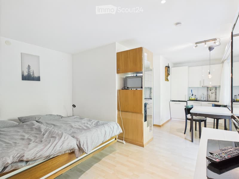 Bedroom: bed, pillow, blanket, white wall, white floor, wooden shelves, small TV, kitchen island, dining table, chair