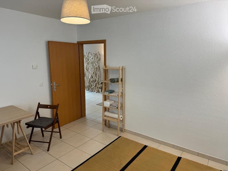 Small empty room with white walls, wooden table, folding chair, wooden shelf, tiled floor, and ceiling lamp.