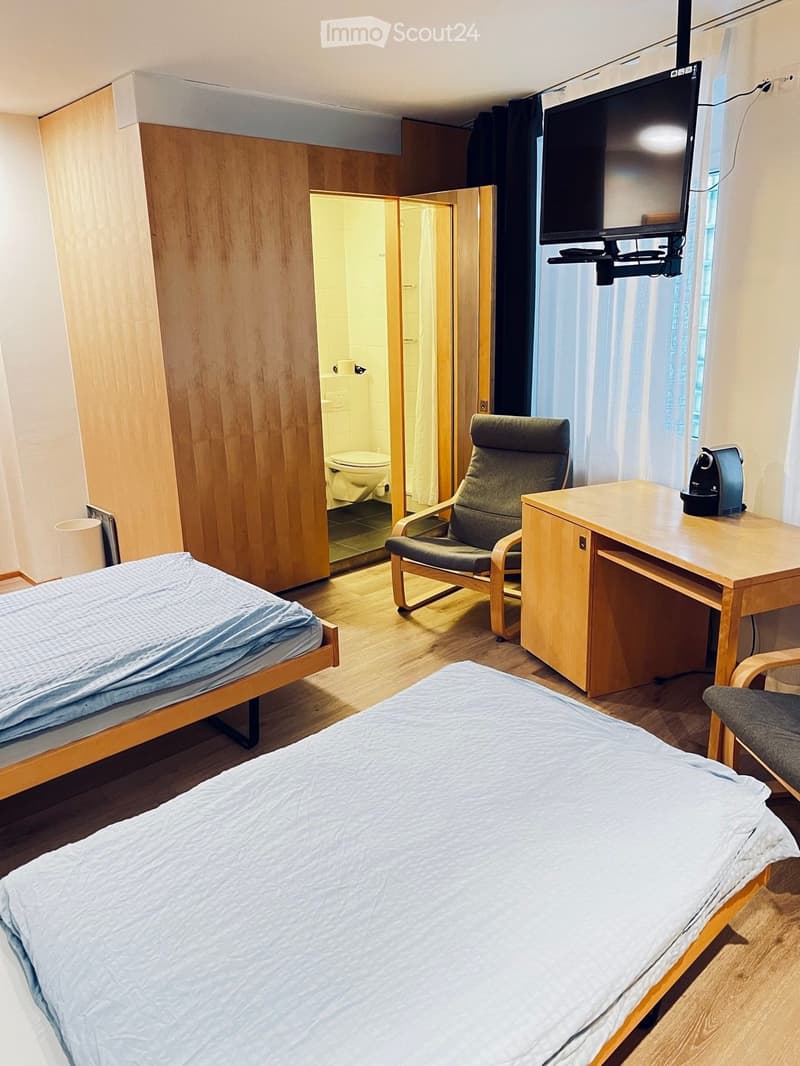 Room with wooden flooring, mounted TV, desk, wooden chair, double bed, wooden cabinet, toilet, waste bin, bed covers, and desk accessories