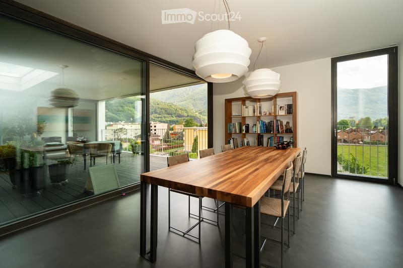 Dining room with large windows, wooden table, chairs, bookshelves, and a balcony with a view of the mountains.