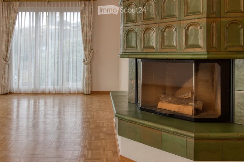 Empty room with wooden parquet flooring, green tiled fireplace, large window with white curtains, wooden floor, light colored walls