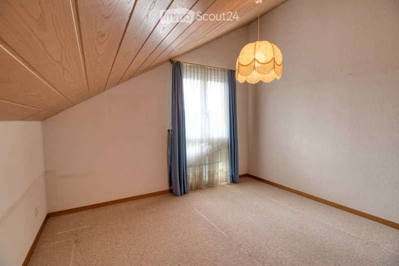 An empty room with wooden ceiling, white walls, a large window, white curtains, a wooden strip at the base of the wall, and a lamp hanging from the ceiling.