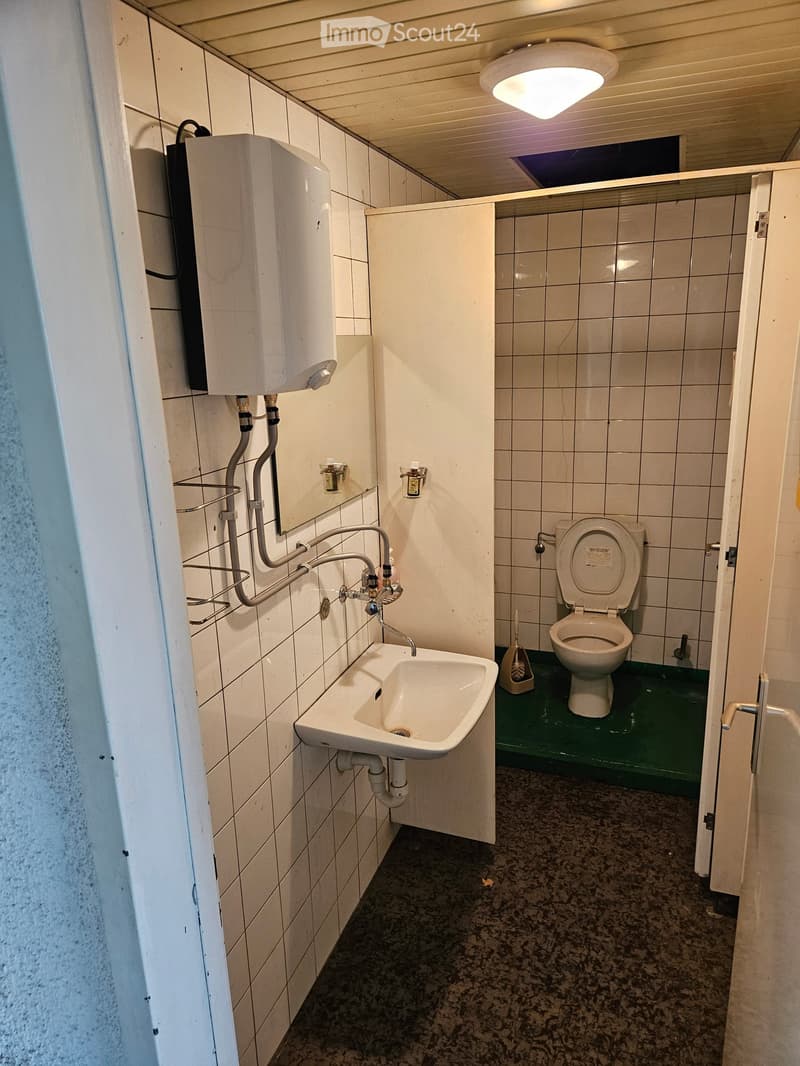 A small bathroom with tiled walls and floors, a sink with faucet, a toilet, a mirror, and a washing machine above the sink.