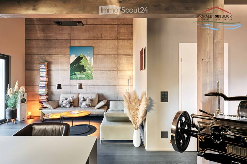 concrete wall, gray couch, white side table, round wood table, gray ottoman, black chair, white vase with dry grass, books, stack, modern art, speaker