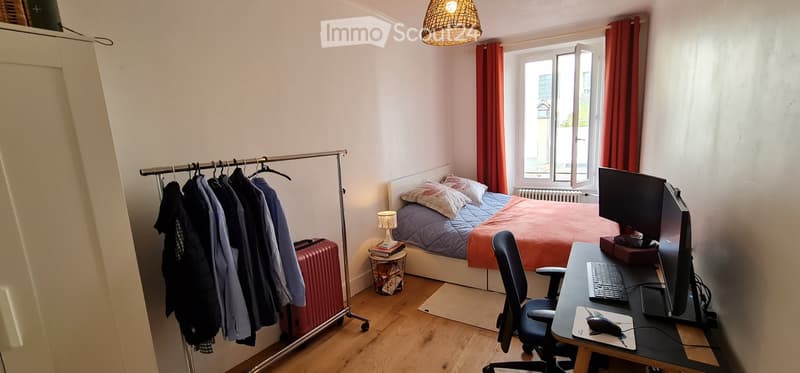 A room with wooden floor, wardrobe, clothes hanger, single bed with pillows and sheets, desk with computer and mouse, chair, table lamp, radiator, open window with curtains