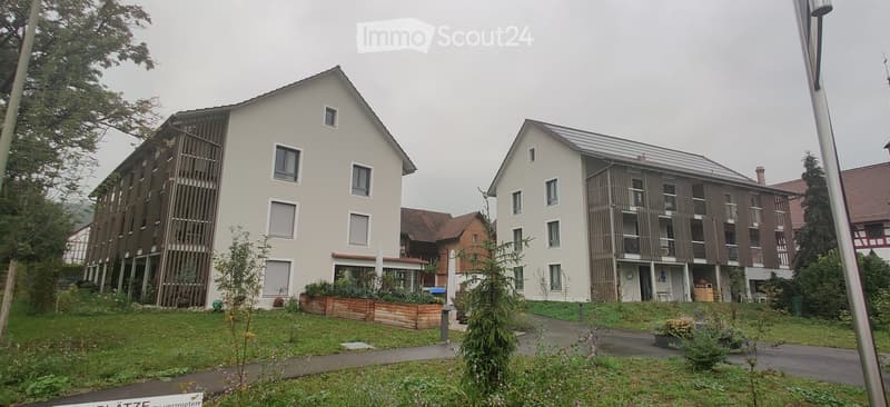 Two residential apartment buildings, front view, solar panels on one, surrounded by trees and grass