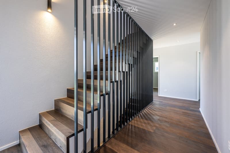 Wooden staircase with a metal handrail, modern lighting design on the wall, wooden flooring, and a minimalist aesthetic.