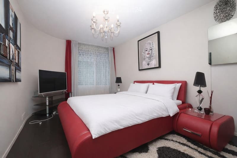 Red bed, white bedding, two nightstands with lamps, TV on a stand, picture frame, mirror, chandelier, black and white rug