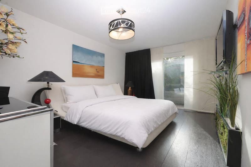 modern bedroom with white bed, white bedding, white walls, black curtains, ceiling lamp, wall mounted TV, artwork, nightstand with lamp, potted plant