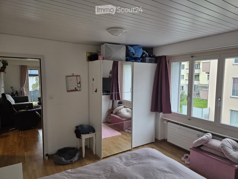 Bedroom with bed, wardrobe, stool, and pink rug. Large windows with curtains. View of another room with couch and plant.