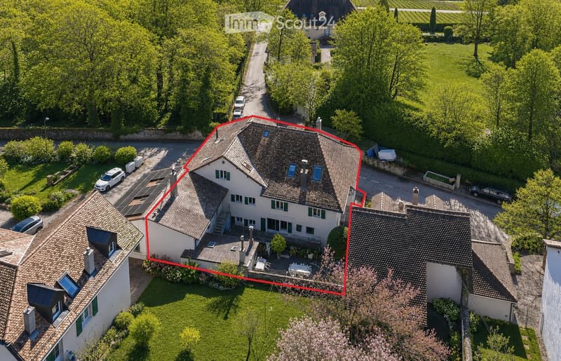 Aerial view of a two-story house with a tiled roof, surrounded by a well-maintained garden and driveway. The house features multiple windows, a small patio area with outdoor furniture, and is situated in a residential neighborhood with other houses and gr