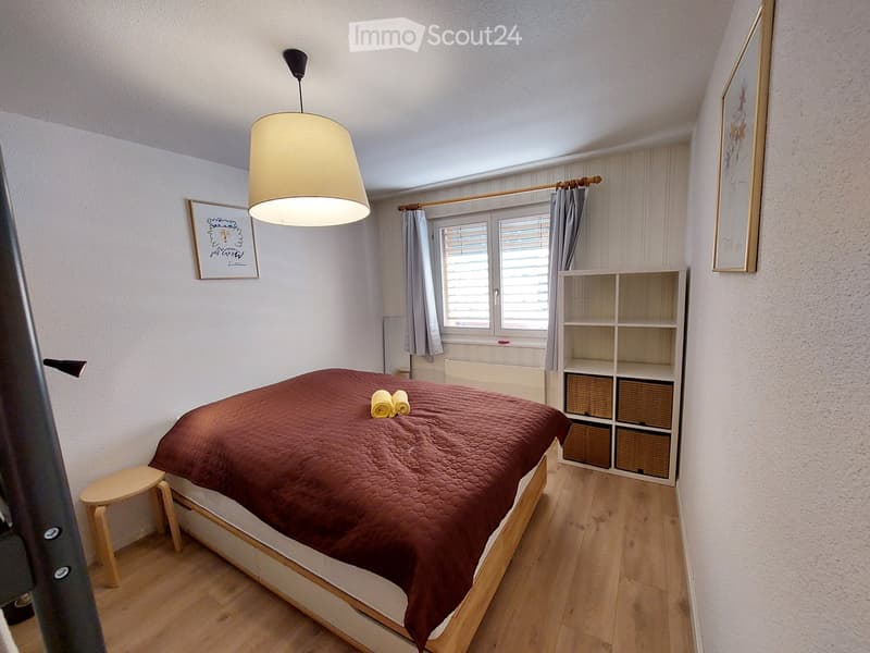 Bedroom with bed, wooden floor, white walls, lamp, window with curtains, stool, shelves, picture frames