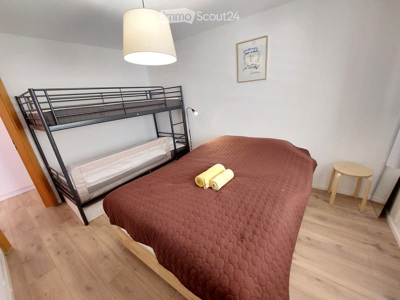 Bedroom with a double bed, bunk bed, wooden floor, white walls, lamp, picture frame, and a stool with towels.