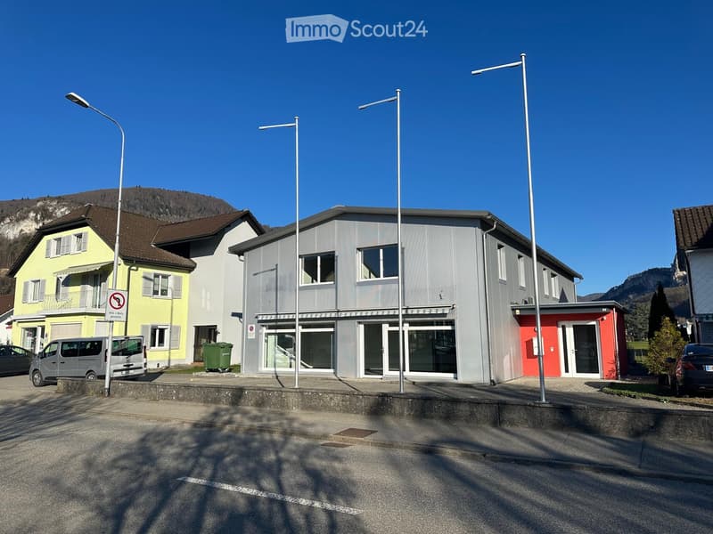 A gray building with a red annex, glass windows, and a red door. There are street lights and a signboard on the left side. A van is parked in front of the building. A trash bin is placed on the side of the road. A car is parked on the right side. A mounta