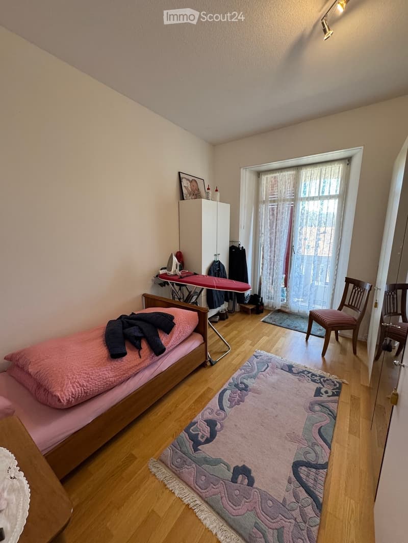 Single bed, wooden floor, rug, chair, ironing board, cabinet, picture frame, window with curtains