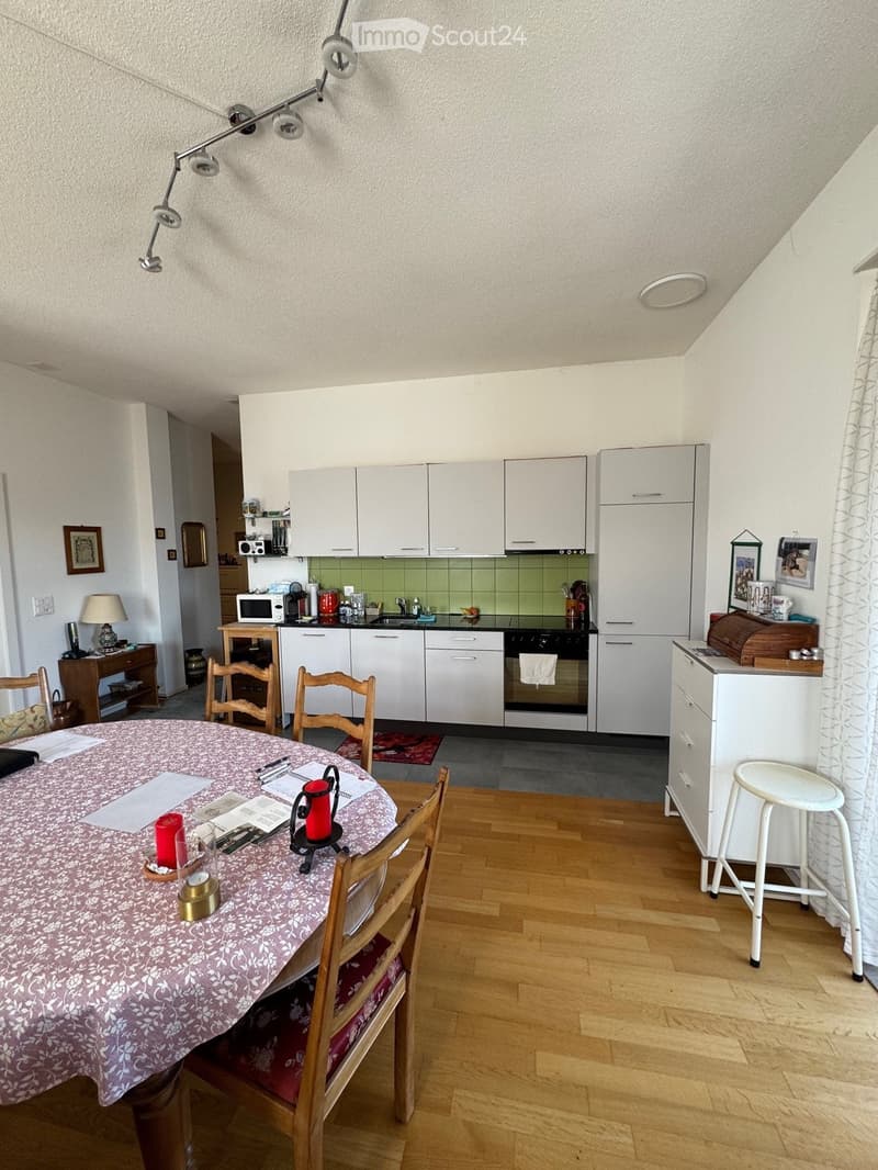 Dining room with a table and chairs, kitchen with white cabinets, green tiles, microwave, oven, and a white stool