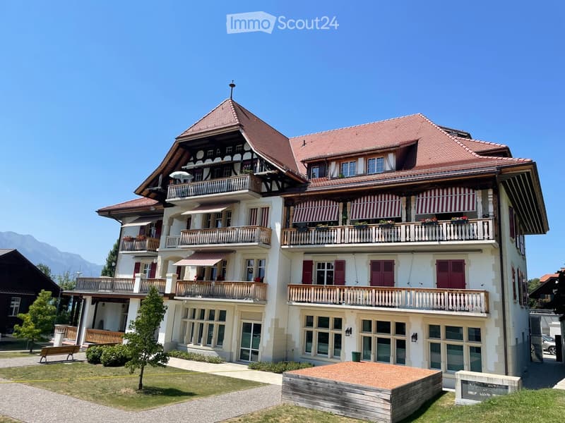 A large, multi-story house with a red-tiled roof, white walls, and multiple balconies adorned with striped awnings and flower boxes. The house features a mix of traditional and modern architectural elements, including wooden beams and large windows.