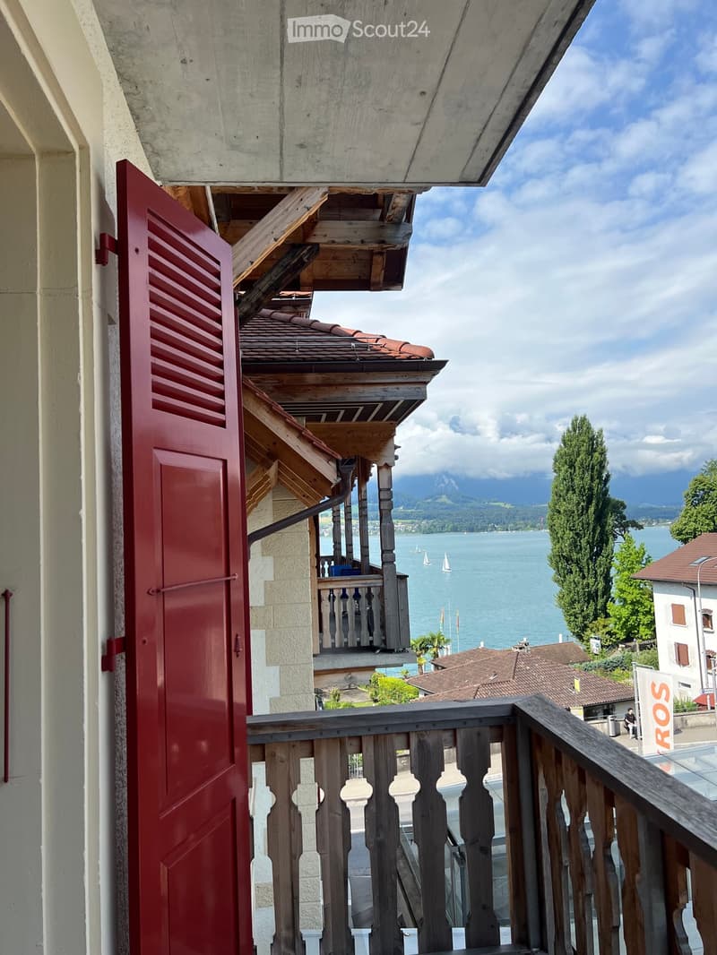 Balcony overlooking a scenic lake with sailboats and distant mountains, featuring wooden railings and a red shuttered door.