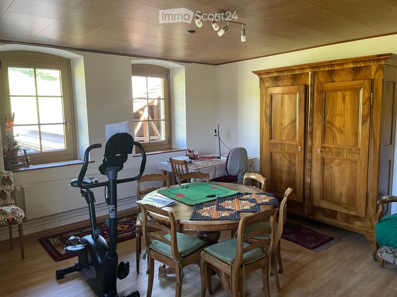 Well-lit room with wooden floors, large windows, exercise bike, dining table with chairs, and a large wooden cabinet.