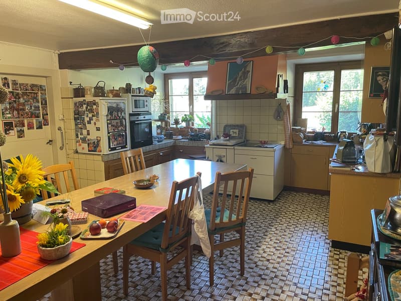 Bright kitchen with a wooden dining table and chairs, refrigerator, microwave, oven, and various kitchen appliances. Decorated with flowers and framed pictures.