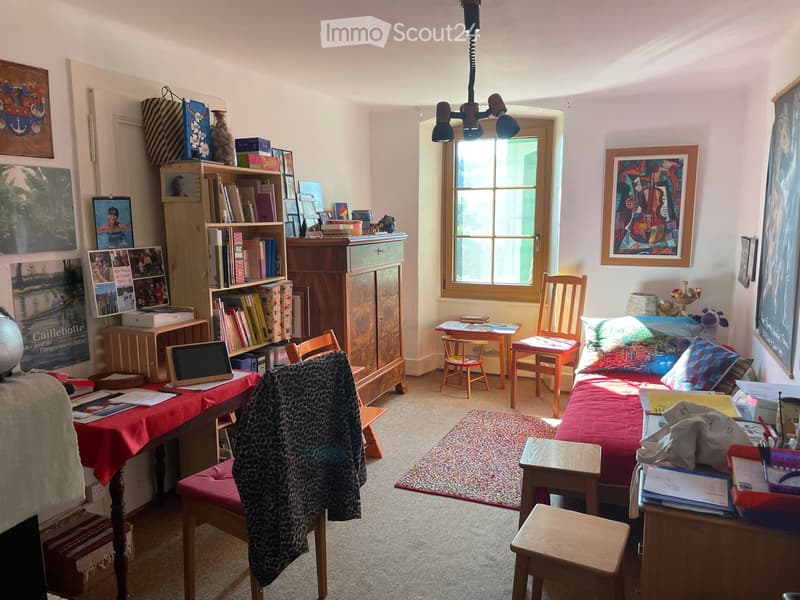 A cozy, well-furnished room with a bed, laptop on a table, bookshelf, and various chairs and stools. The room features artwork on the walls and a window letting in natural light.