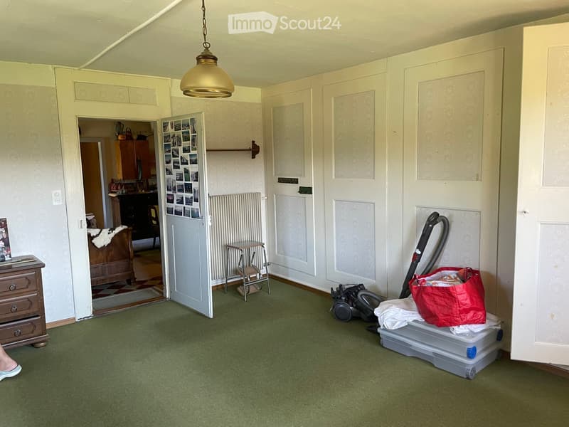 Empty room with green carpet, white walls, and a hanging light fixture. A vacuum cleaner, a red bag, and some storage boxes are placed near the door.