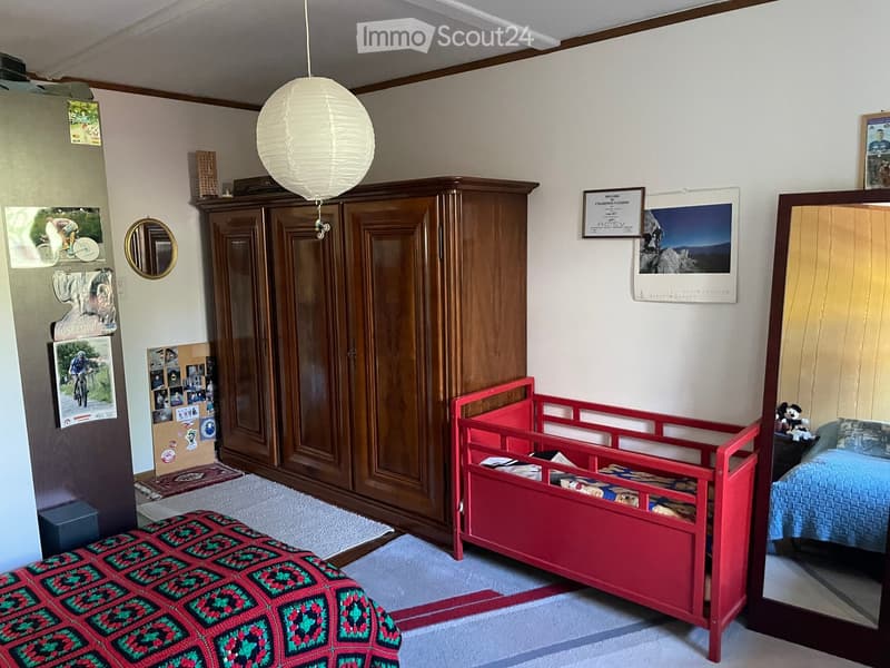 A cozy bedroom with a large wooden wardrobe, a red toy bed, a small bed with a blue blanket, a round mirror, and various decorations including a calendar and picture frames.