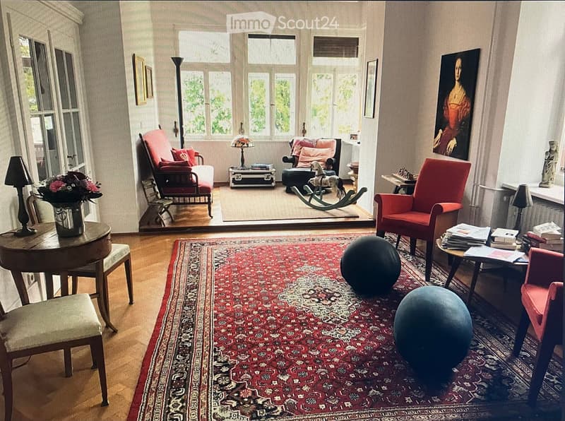 large living room, wooden floor, large windows, red couch, red armchairs, table, rug, lamp, books, flowers