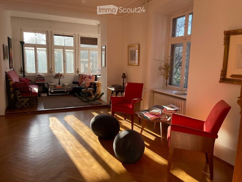 Living room with wooden floor, red chairs, coffee table, chess board, radiator, windows, picture frames, rocking chair, lamp, books, and plants