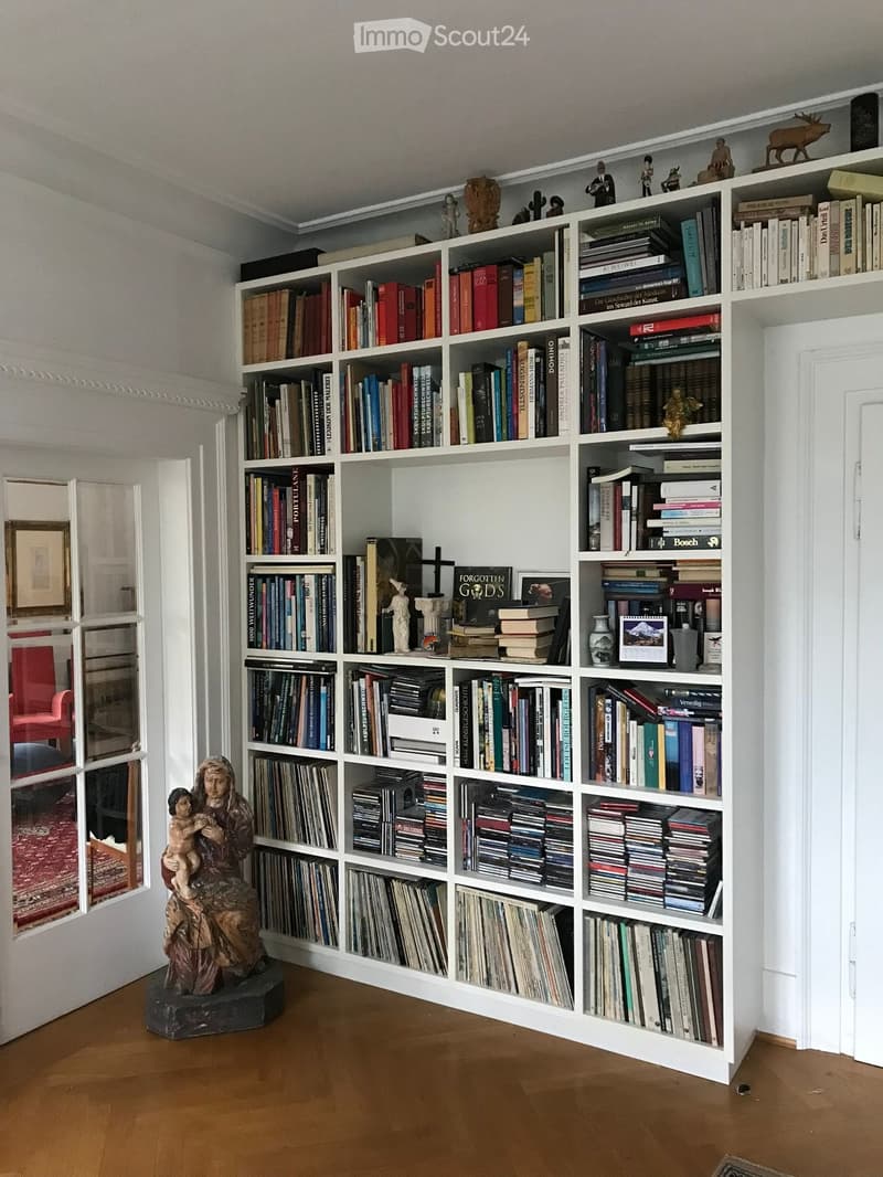 A room with a large white bookshelf filled with books, CDs, and decorative items. A wooden statue is on the floor in front of the bookshelf. The room has a wooden floor and white walls.