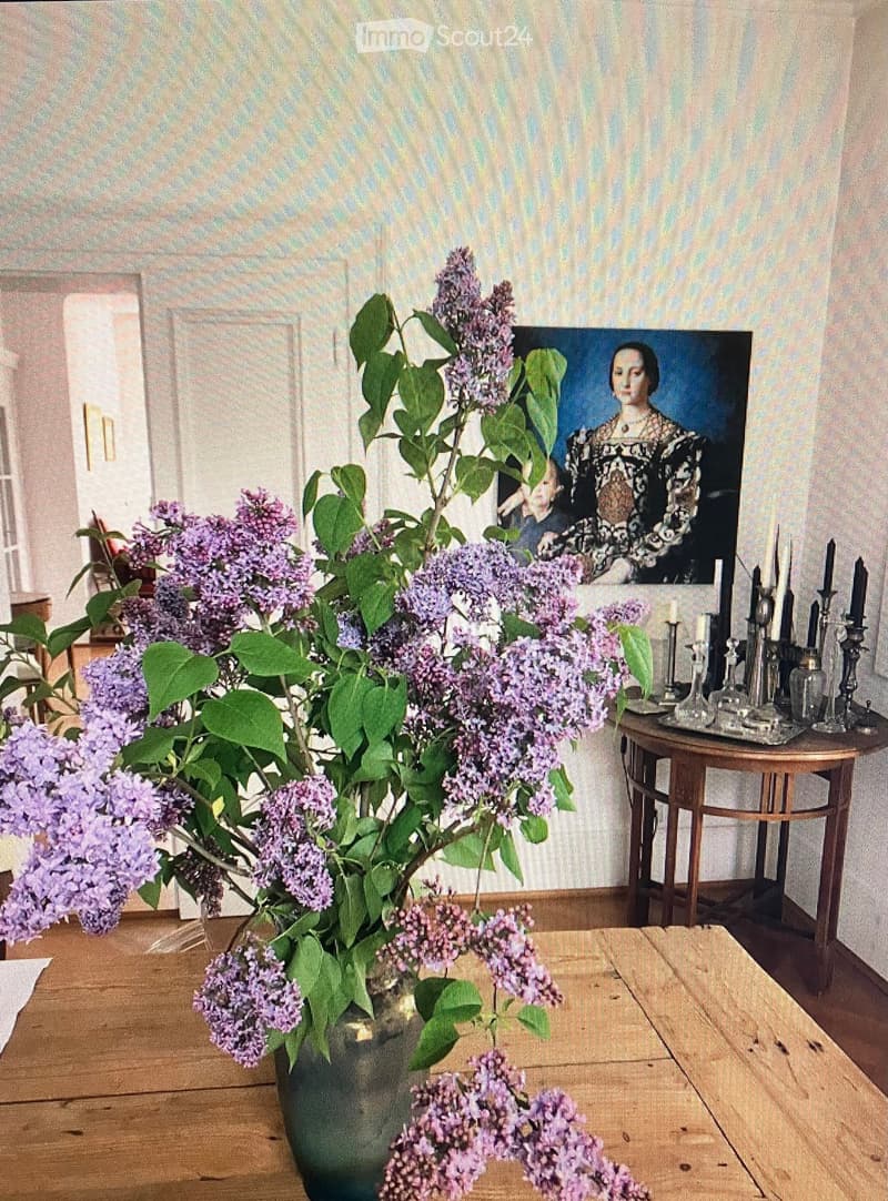 A wooden table with a vase of lilacs, a small table with candles, a painting of a woman, and a patterned wall.