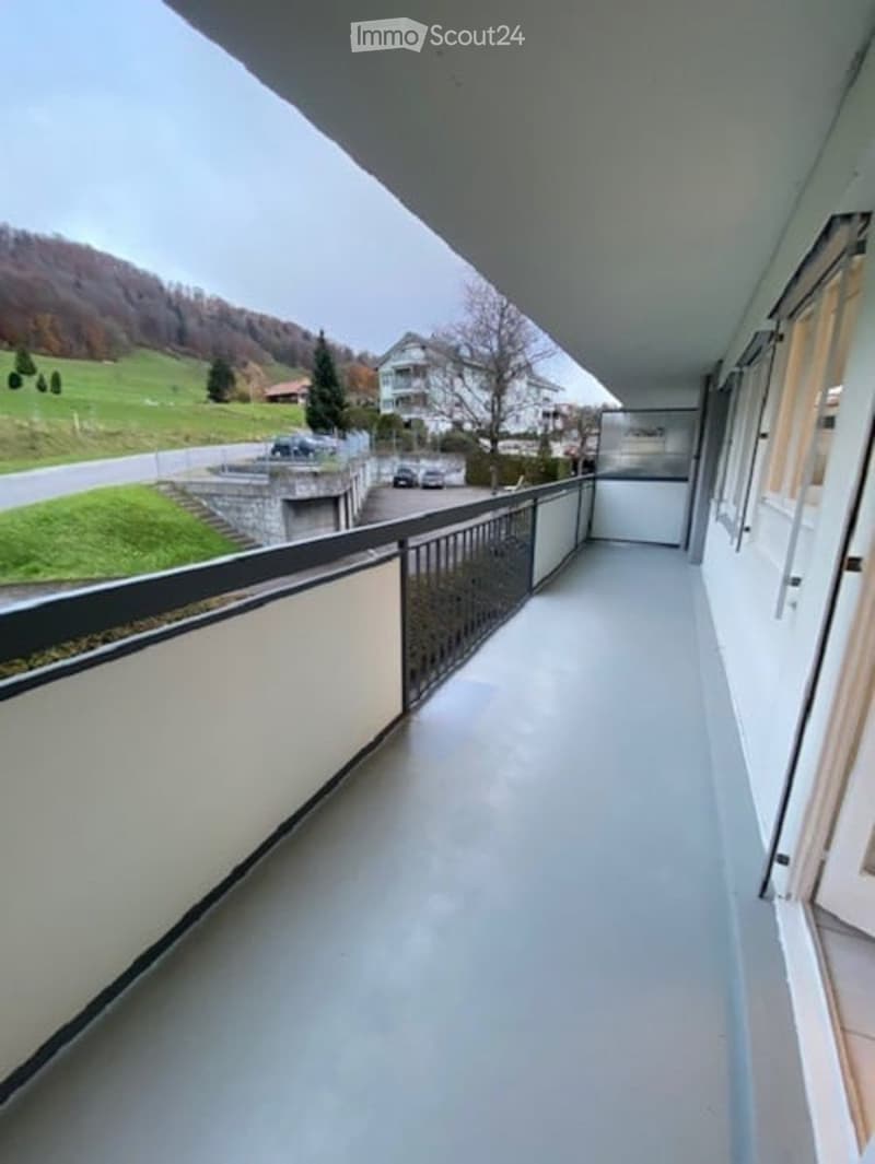Balcony with railings, white floor, white walls, view of a grassy hill and houses