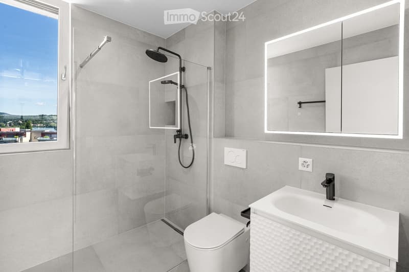 Modern bathroom with grey tiles, a white toilet, a sink with a black faucet, a large mirror with built-in lighting, and a shower with a handheld sprayer.