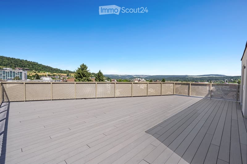 Wooden deck with a modern woven railing, overlooking a scenic landscape with trees and distant buildings.
