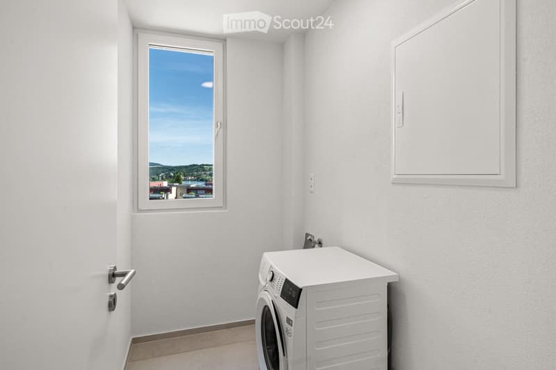 Empty room with white walls, a window showing a scenic view, a door, and a private washing machine installed against the wall.