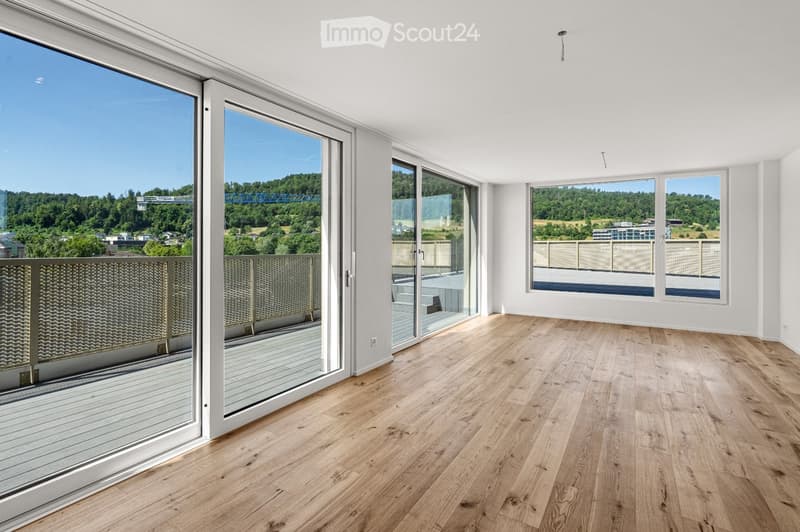 Bright, unfurnished room with large windows and wooden flooring, offering scenic views of greenery and a bridge.
