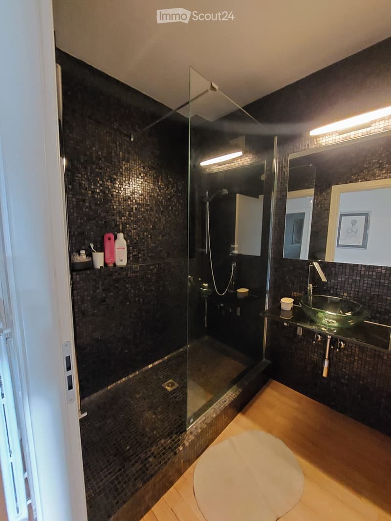 Modern bathroom with glass shower, black tiled walls, wooden floor, glass sink, mirror, and a shelf with toiletries